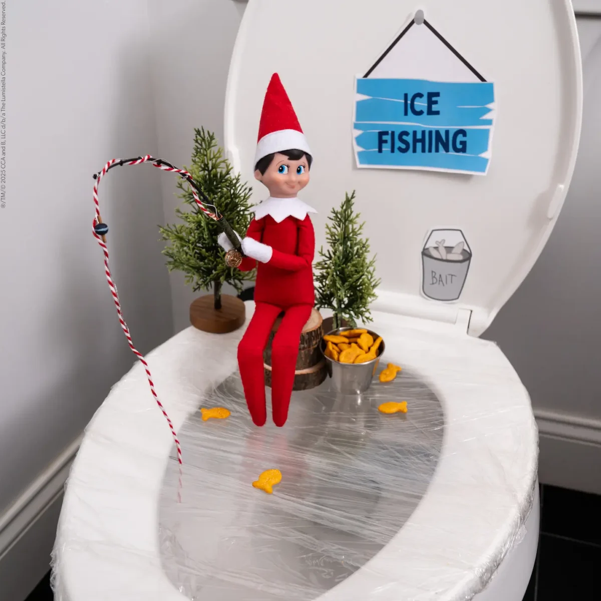 The Elf on the Shelf sits on a toilet wrapped with plastic wrap as with a sign that says Ice Fishing