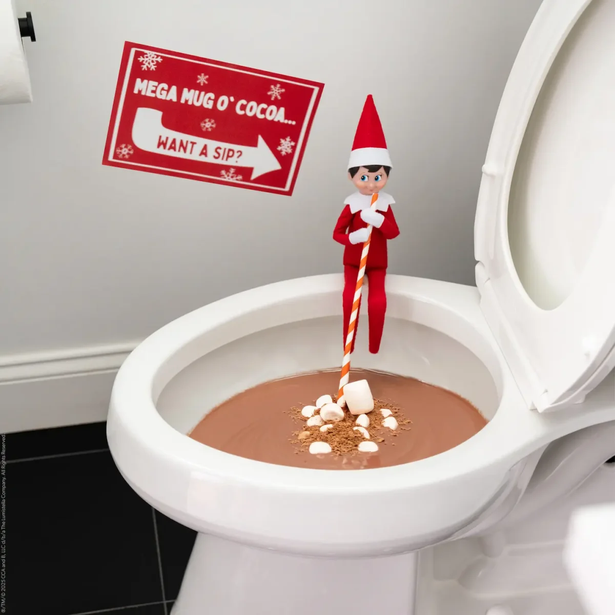 A Scout Elf holds a long straw while sitting on the side of a toilet filled with cocoa and marshmallows, a sign on the wall says "Want a sip?"