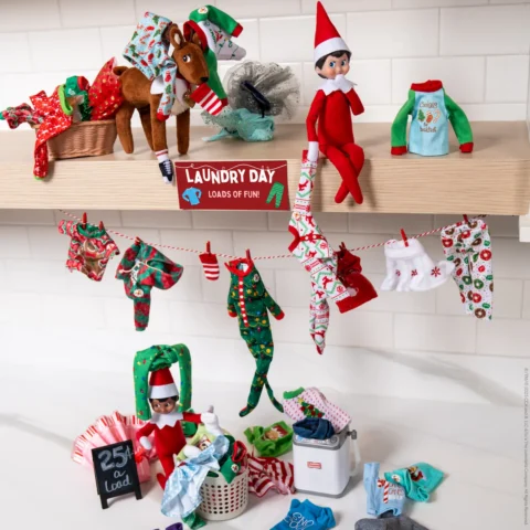 Scout Elf on the Shelf sitting beside a mini clothesline with a “Laundry Day” sign and tiny elf-sized clothing in the laundry room.