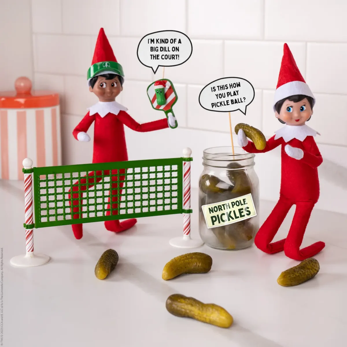 Two Elf on the Shelf Scout Elves stand on either side of a net holding paddles, one of which has a pickle resting on it) and a sign that says, "Is this how you play pickleball?"