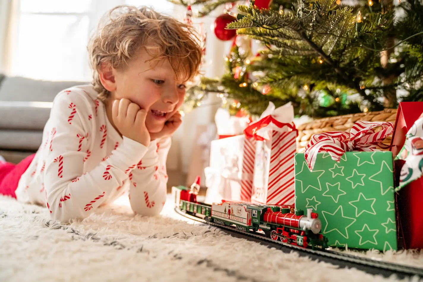 A young boy plays with a train underneath a Christmas tree with twinkling lights. The miniature Elf on the Shelf rides in the back of the train