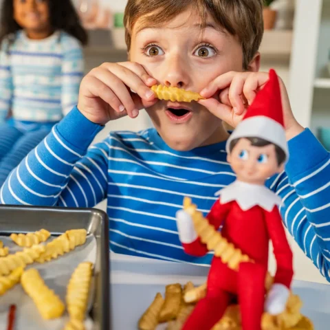 A boy holds a fry up like a mustache while standing behind The Elf on the Shelf