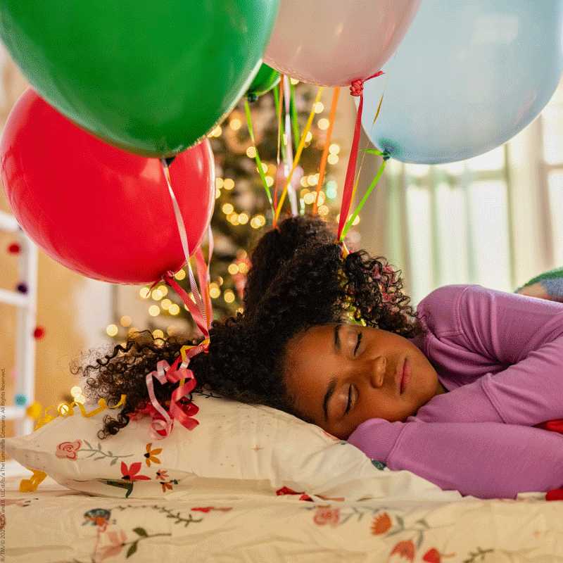 GIF showing a girl sleeping, then waking up to see balloons have been tied to her hair, then sitting up in delight.