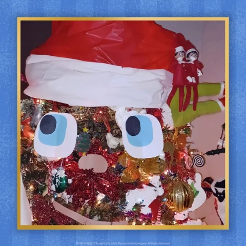 A Christmas tree decorated as a large Elf on the Shelf with two small elves sitting on the brim of the tree's elf hat