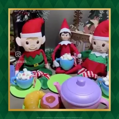 The Elf on the Shelf holds a cup of marshmallows while sitting between two stuffed dolls 