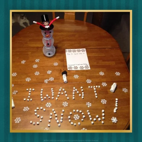 The Elf on the Shelf sits on a table where he has spelled out I Want Snow using marshmallows