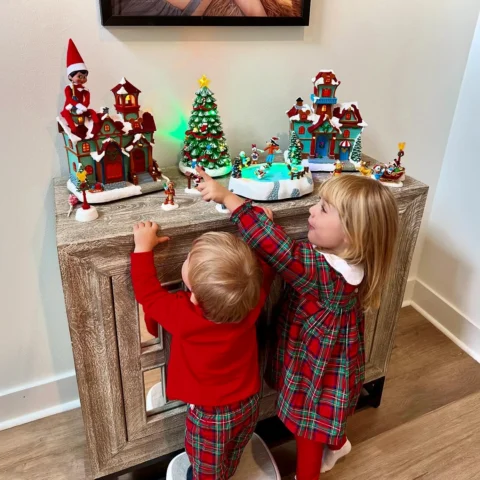 A pair of children are smiling and pointing to their posed Scout Elf atop a Christmas village decoration rooftop
