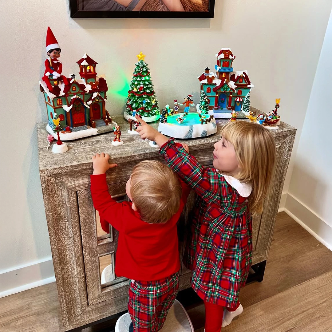 A pair of children are smiling and pointing to their posed Scout Elf atop a Christmas village decoration rooftop