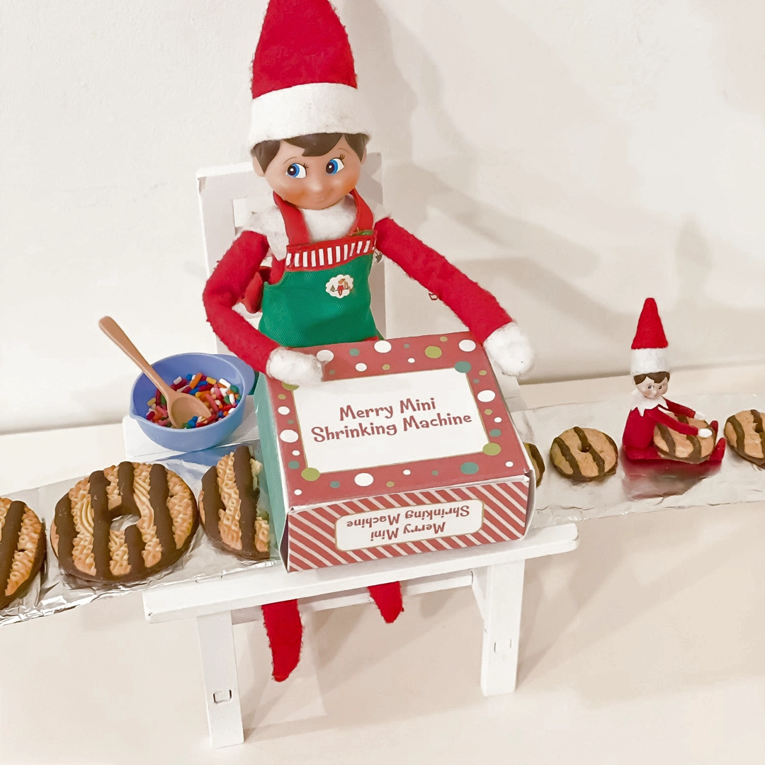 A Scout Elf sits in front of a makeshift "Merry Mini Shrinking Machine" with a homemade conveyor belt of Fudge Round style cookies entering into the machine, followed by smaller versions, including a shrunken ScoutElf!