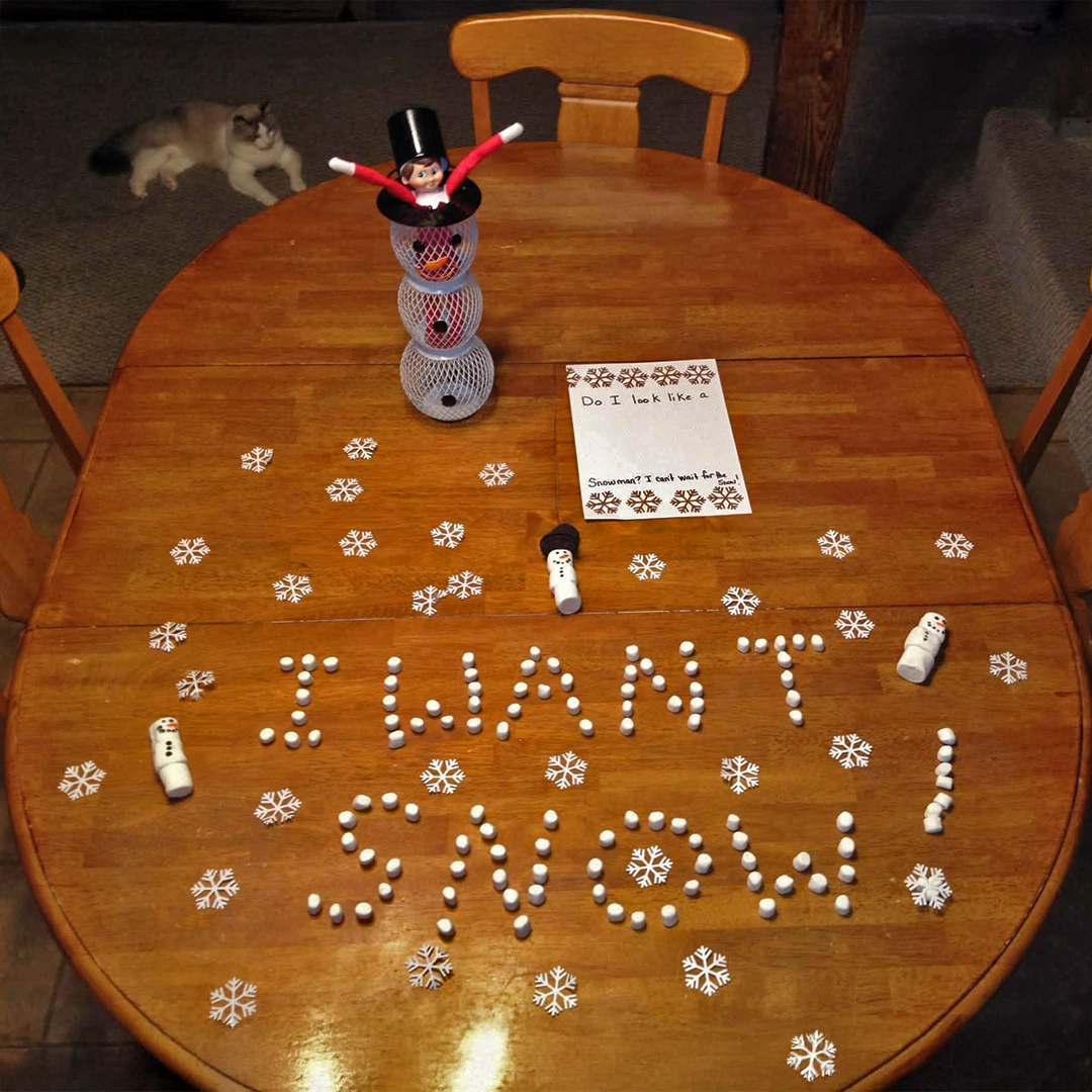A Scout Elf is posed on a dining table in a wire snowman container. Strewn about the table are paper snowflakes, a note reading, "Do I look like a snowman? I can't wait for the snow!" There are a few marshmallow snowmen on the table, as well as "I WANT SNOW!" spelled out in smaller marshmallows.