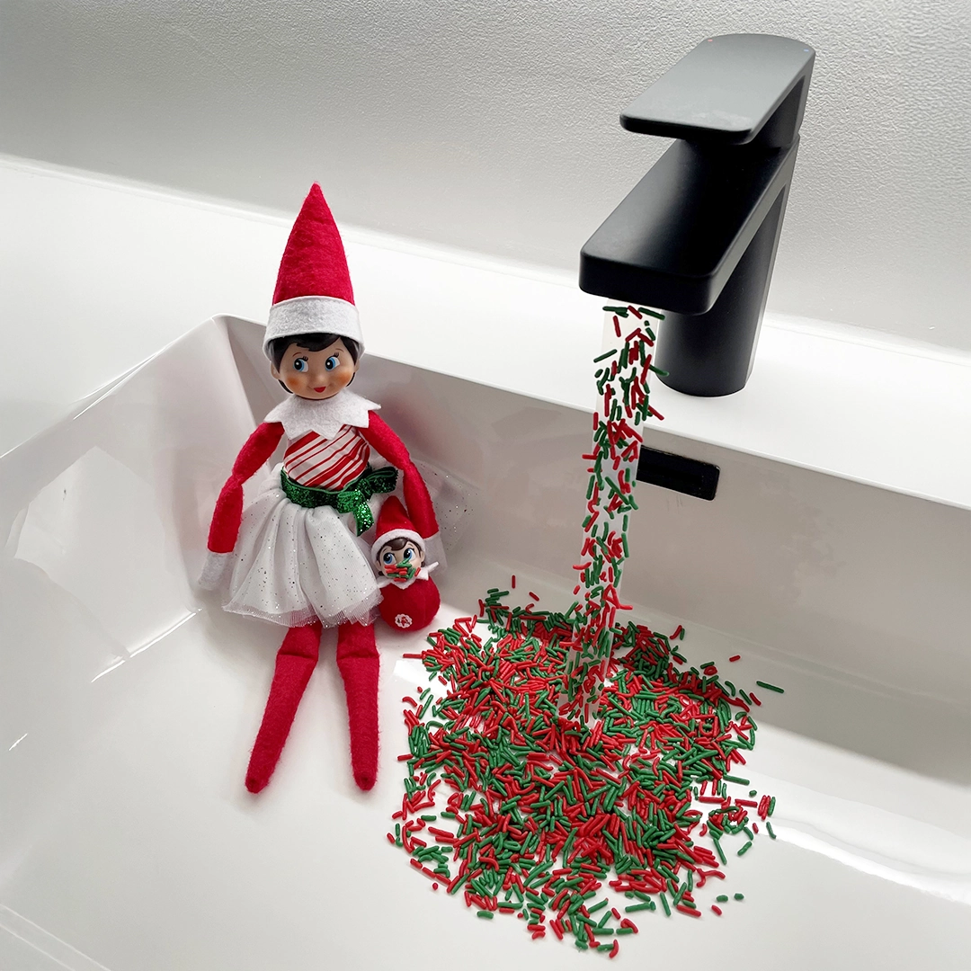 A Scout Elf sits in a sink basin with a Frost Pip elf baby, next to a wondrous fountain of red and green sprinkles! The sprinkles are stuck to transparent tape, giving them the effect of flowing from the faucet.