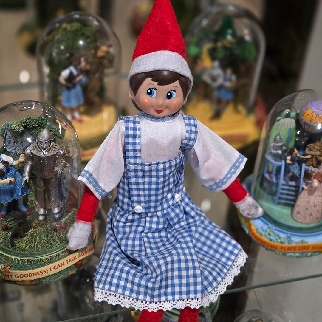 A girl Scout Elf is dressed in a Dorothy style dress, amidst a collection of Wizard of Oz snow globes!