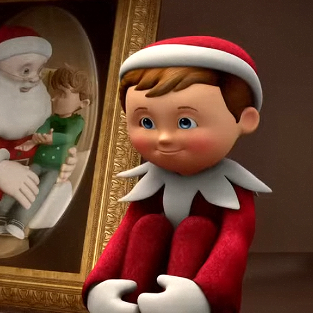 A still of Chippey from the animated special, "An Elf's Story: The Elf on the Shelf". This particular scene shows Chippey sitting on the shelf in the classic pose. Next to him is a framed photo, further illustrating that he is seated on a shelf.