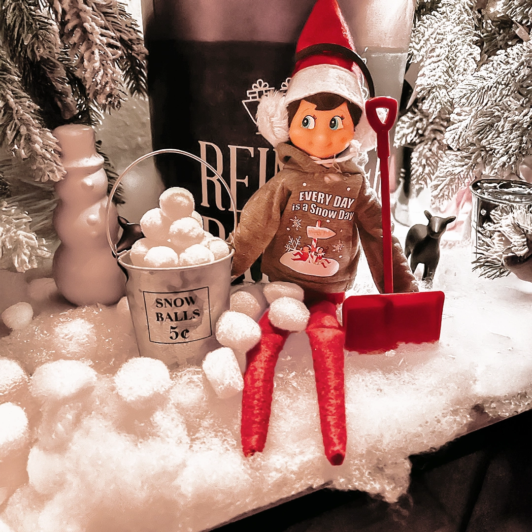 A Scout Elf is seated amongst snowy winterland decor, with a pail full of snowballs and a snow shovel. He is sporting a pair of ear muffs and a snow shovel, as well as the Claus Couture "Every Day is a Snow Day" hoodie.