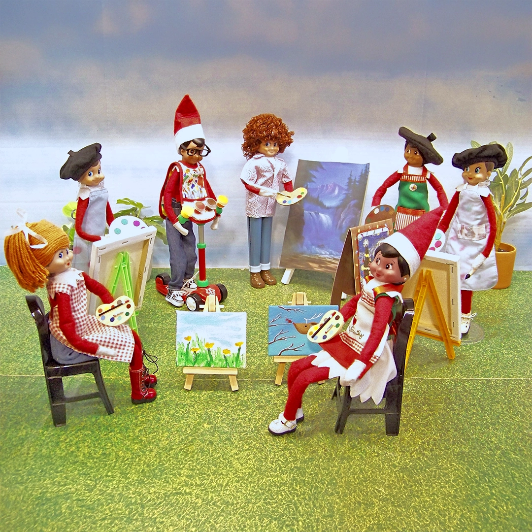 A group of Scout Elves are posed together in a circle among miniature canvases, with painting pallets, chairs and easels. One of the Scout Elves slightly resembles Bob Ross. They are seated in an area with print outs of grass and a brilliant, blue sky.