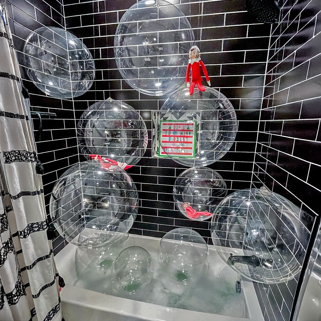 A Scout Elf is seemingly floating atop a large bubble, which appears to be a clear beach ball. Two more can be seen inside different large "bubbles". These are positioned in a bathroom over a filled tub of bubble bath. The bath tub has a black subway tile surround, making for a striking contrast with the bubbles and Scout Elves.