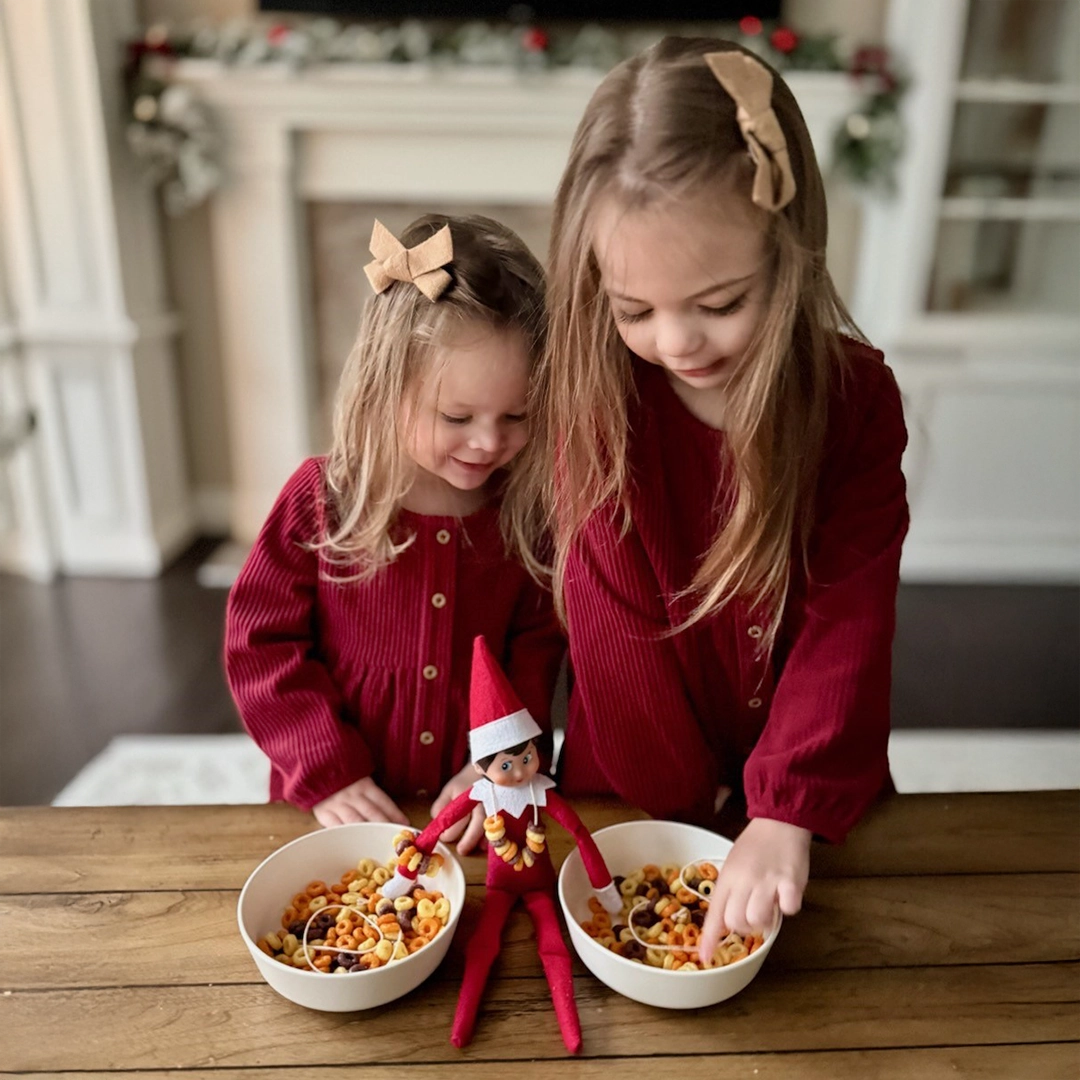 A pair of young girls adorned in matching, burgundy Christmas dresses and tan bows are rifling through a pair of cereal bowls filled with "O" shaped cereal, each to make a set of "cereal bead bestie bracelets". Their Scout Elf is seated in between the bowls, wearing a necklace made in the same fashion.
