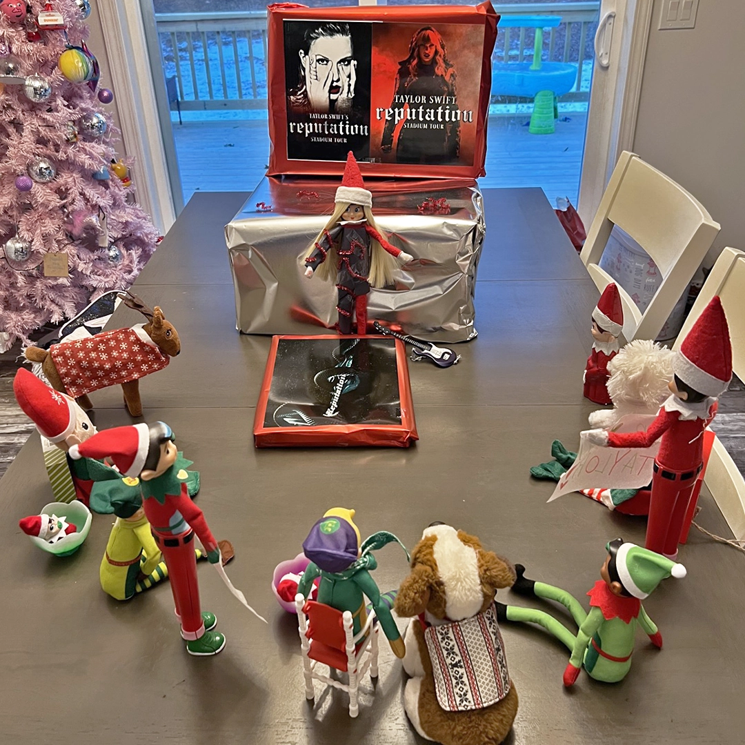 A crowd of Scout Elves and Santaverse favorites, like the Elf Mates Chef, Cobbler, and Toy Maker as well as the Elf Pets Saint Bernard and Reindeer gather for an impromptu Taylor Swift cover band concert starring Taylor's elf doppelgänger. Her stage is assembled from wrapped gifts adorned with print-outs of two Reputation tour posters.