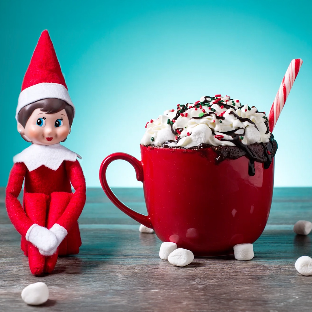 A girl Scout Elf is seated next to a red coffee mug filled with a Cocoa Cup Cake, a candy cane and chocolate syrup which is dripping over the edge of the mug. This pose is a still from the No-Mess Cocoa Cup Cake recipe video on YouTube. The background is a backlit light blue backdrop that has a gradient effect.