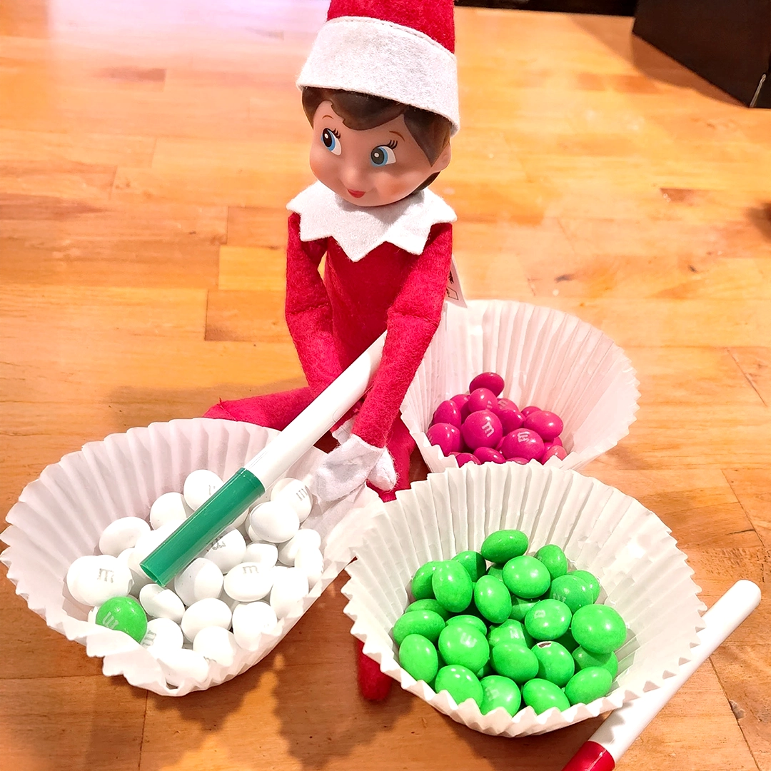 A girl Scout Elf is posing seated on a kitchen table with 3 cupcake liners, each filled with their own Christmas colored M&Ms candies. One is red, the other light green, and the last is white. The Scout Elf also has a red and green marker. The white M&Ms group has a single green M&M, alluding to the Scout Elf coloring them all!