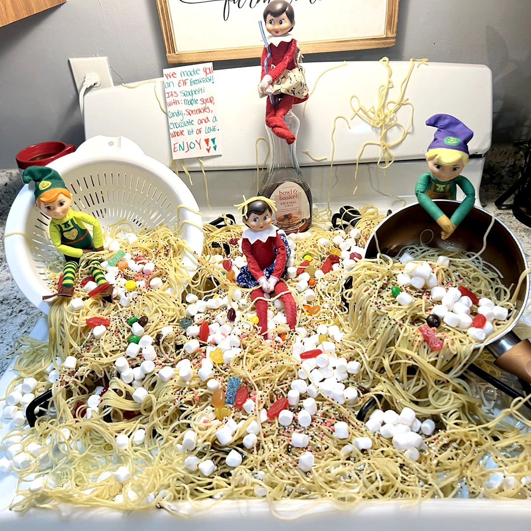 A pair of Scout Elves and Elf Mates are posed on the stovetop in a kitchen amongst a huge mess of spaghetti noodles, sprinkles, Swedish Fish candies, Sour Patch Kids Candies, marshmallows and presumably syrup with a note that reads, "We made you an elf breakfast! It's spaghetti with: maple syrup, candy, sprinkles, chocolate, and a whole lot of love. Enjoy!!"