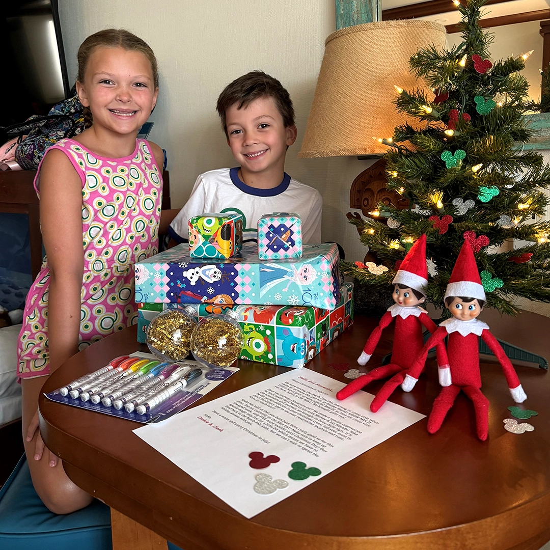 A pair of children pose with a miniature Christmas tree, a stack of presents and an ornament craft, along with a note from their Scout Elves, who are also seated on the table holding the other elements of the photo. They appear to be in a hotel on a family Disney World trip.