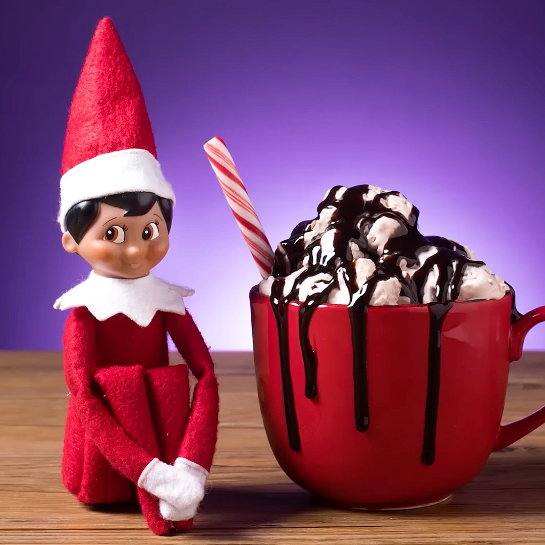 A boy Scout Elf is seated next to a red coffee mug with ice cream, a candy cane and chocolate syrup which is dripping over the edge of the mug. This pose is a still from the Hot Cocoa Ice Cream recipe video on YouTube. The background is a backlit purple backdrop that has a gradient effect.