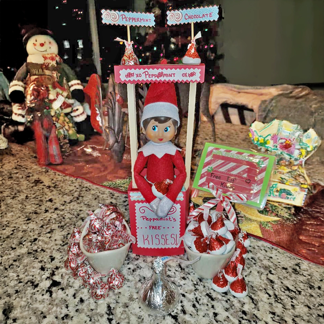 A female Scout Elf is manning a Kissing Booth with Hershey's Kisses candies. Christmas decor and a lit tree are seen in the background.