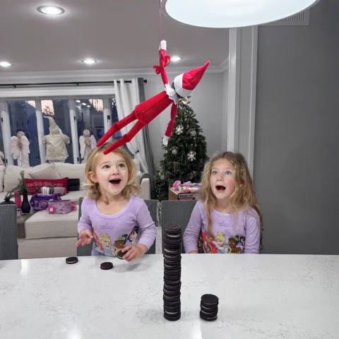 A Scout Elf repels from a pendant light in the kitchen, hovering over a stack of Oreo style cookies. A pair of girls look onto the scene in awe, amazement and joy!