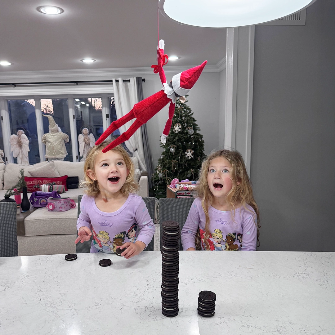 A Scout Elf repels from a pendant light in the kitchen, hovering over a stack of Oreo style cookies. A pair of girls look onto the scene in awe, amazement and joy!