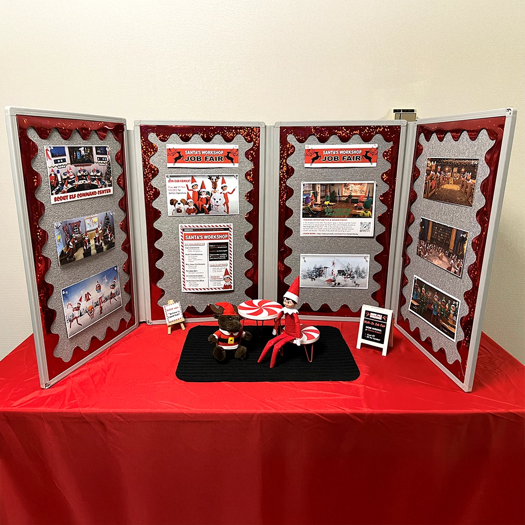 A Scout Elf conducts and interview for one of Santa's newest potential helpers at Santa's Workshop Job Fair events. There is a 4-panel job display board surrounding them, as well as a few signs advertising these newly open positions.
