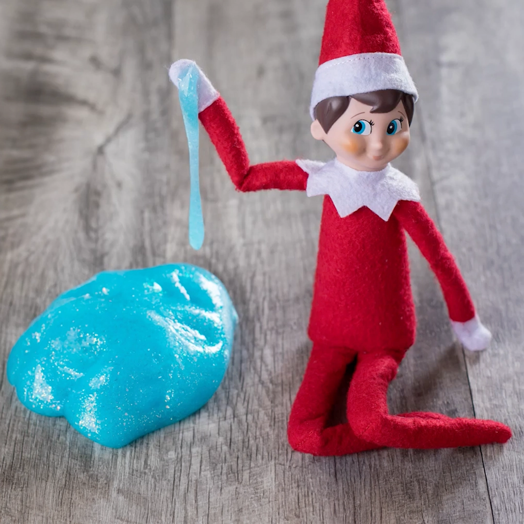 A boy Scout Elf is posing next to a glob of North Pole Glitter Slime. He is seated on the floor. Some of the slime dangles from his raised hand. This still is from the How to Make North Pole Glitter Slime video on YouTube.
