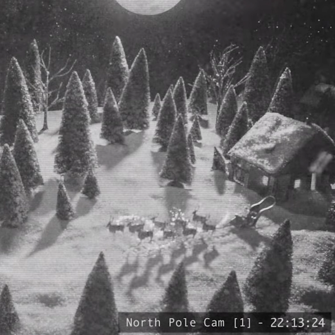 A still from a security camera showing a wintery snowscape with the moon overhead and a snow-topped building. The focus of the video still is of Santa's Sleigh and 8 tiny reindeer. In the bottom right-hand portion of the screen reads "NORTH POLE CAM [1] - 22:13:24" in true security footage fashion.