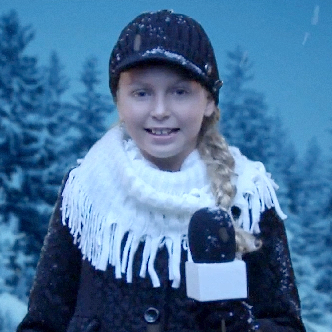 A young girl reporter is reporting from the field in a snowy, winter wonderland. She wears a scarf and hat, while holding a mircrophone. The still is from Reindeer News Network: Will Santa's Sleigh Fly?