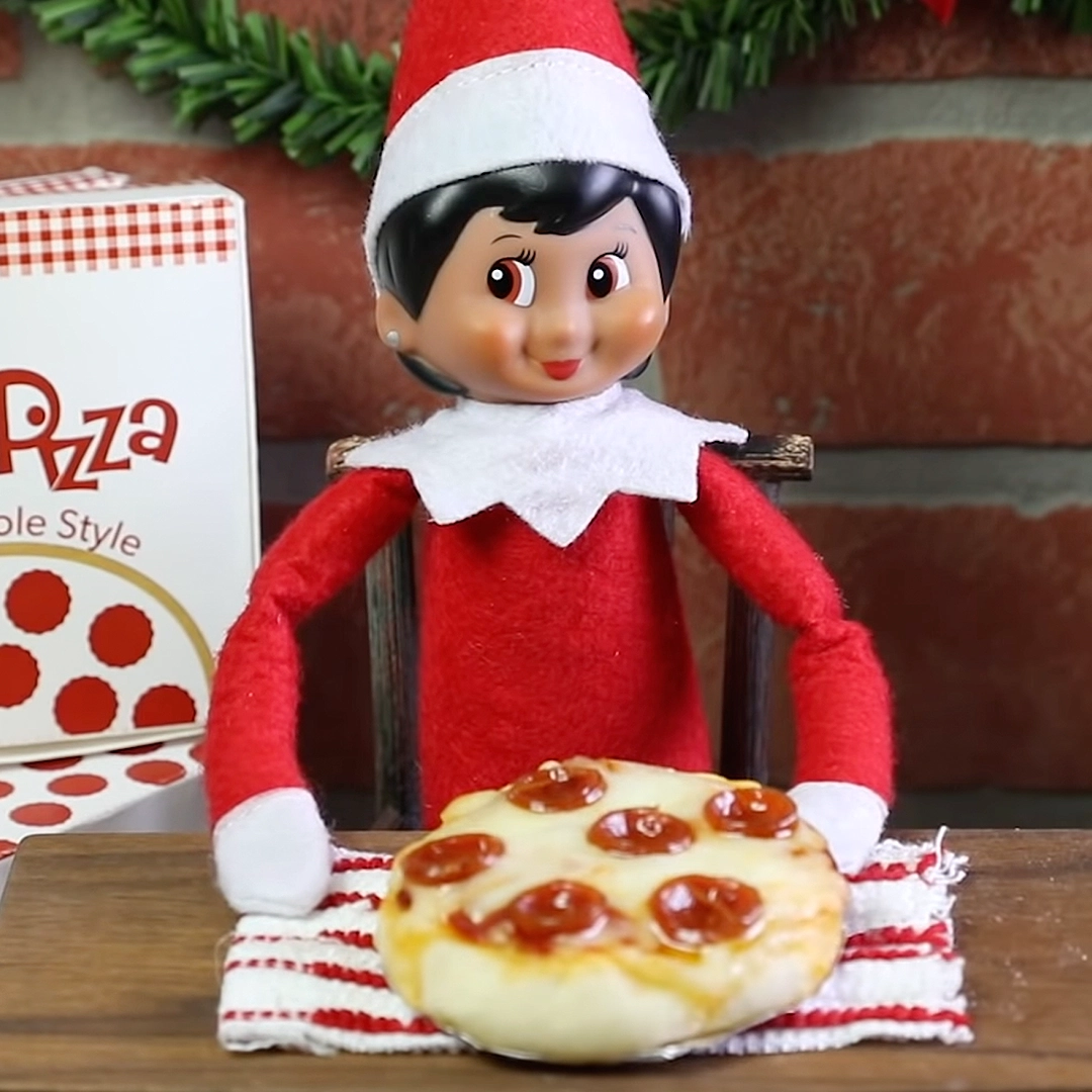 A girl Scout Elf is posing with a mini pepperoni pizza, with a pizza box seen off to the left-hand corner of the image. She is seated at a table with the pizza and a brick wall with a garland strand behind her.