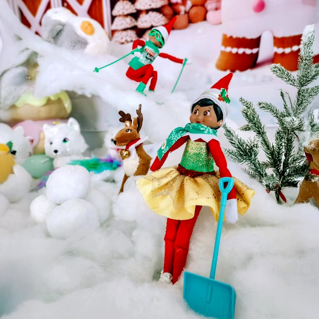 A pair of Scout Elves are staged in a snowy scene, along with Noorah and an Elf Pets Reindeer. The boy Scout Elf is shredding the slopes on his skis, while the girl Scout Elf is looking festive and shoveling snow!