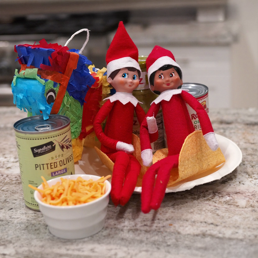 A pair of Scout Elves are seated in taco shells, with all the taco trimmings. They are also amongst a piñata! The scene is staged on a kitchen counter, just in time for taco night!