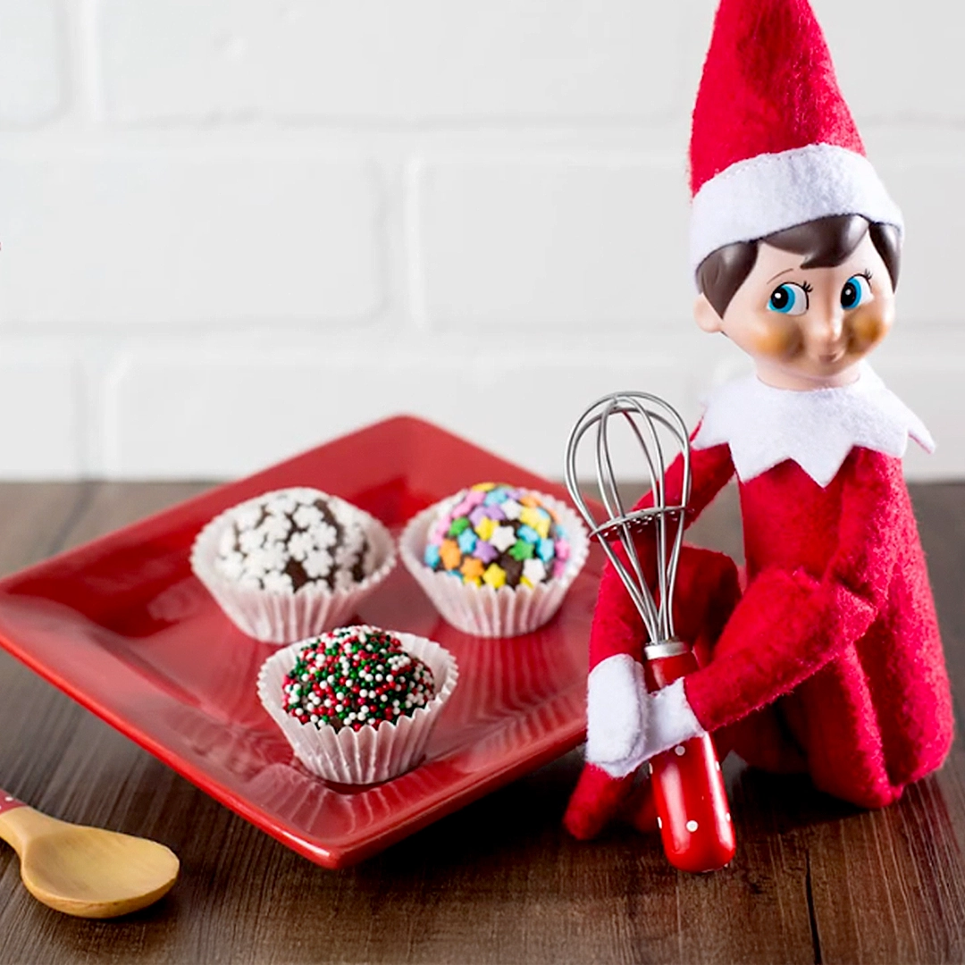 A Scout Elf holds a wisk seated next to a plate of decorated, chocolate truffles from the Tasty Chocolate Truffles Recipe video on YouTube.