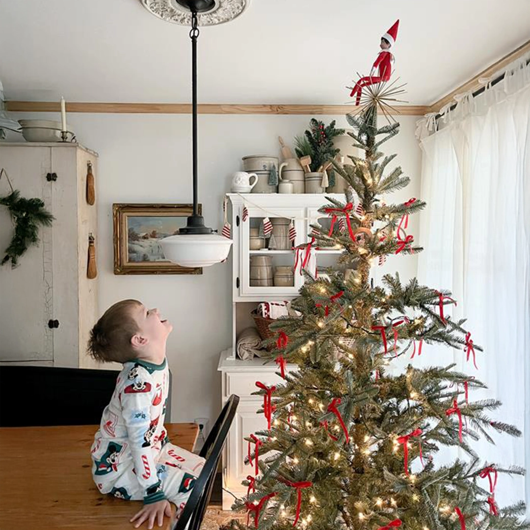 A young boy is seated on his kitchen table looking up with joy and laughter at his Scout Elf, who is seated up high on the family's Christmas tree topper!