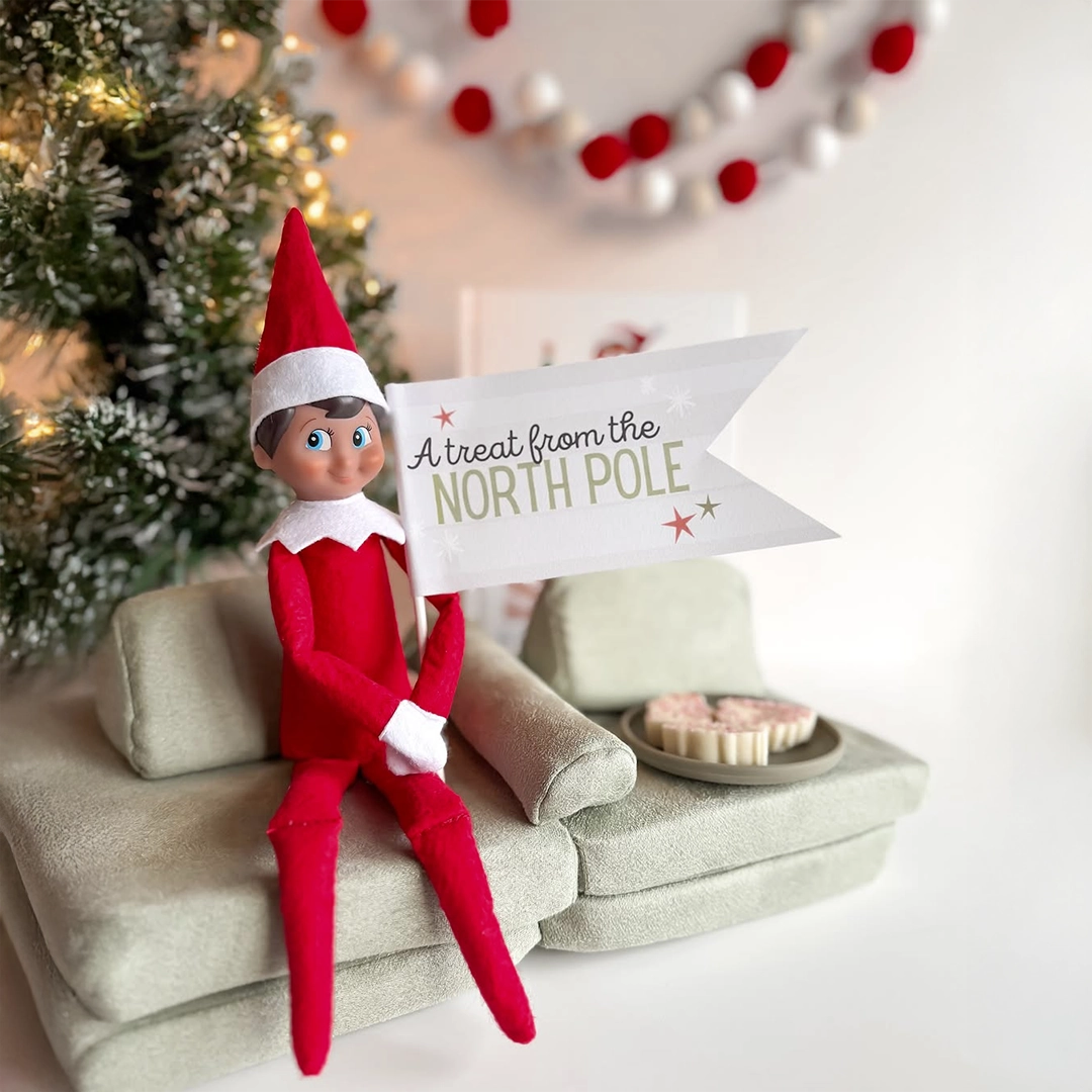 A boy Scout Elf holds a sign reading, "A treat from the North Pole", while seated on a rather comfy but miniature sofa. In the seat next to him are tiny treats from the North Pole! He is seated amongst Christmas decor on what appears to be a shelf.