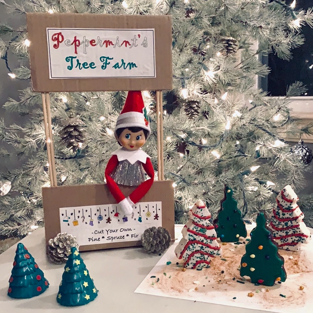 A girl Scout Elf donning the tiny tinsel dress from Claus Couture mans Peppermint's Tree Farm booth, where you cut your own tree. Also shown are a few ceramic conifer tree decor pieces, a pair of pine cones and Little Debbie Christmas Tree cake snacks!