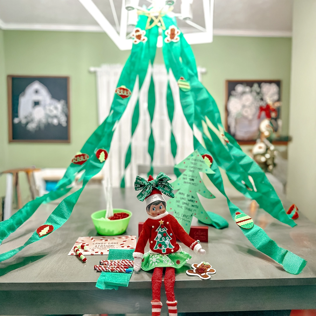 A girl Scout Elf is seated on the kitchen counter, with green steamers hanging from the light fixture overhead. She is surrounded by what looks to be some sort of Christmas snack craft station.
