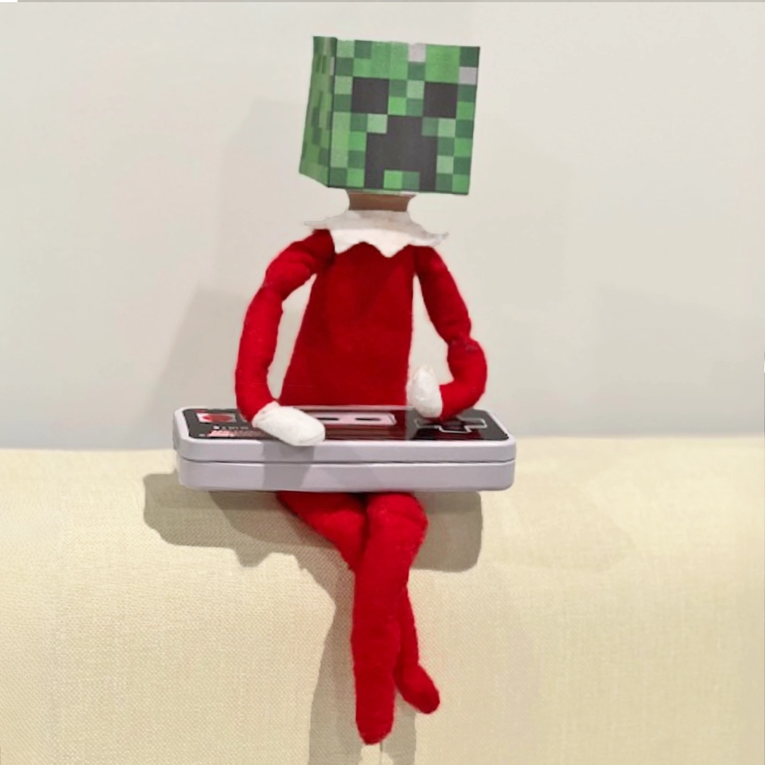 A boy Scout Elf® is seated atop the back of a light upholstered sofa holding a vintage NES controller while wearing a makeshift Minecraft Creeper mask.