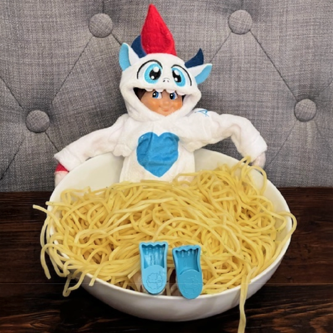 A boy Scout Elf® sporting the Claus Couture Collection® Yeti Hoodie Set relaxes in a bowl of spaghetti noodles with his arms resting along the top of the bowl. The bowl is situated on a dark brown, wooden tabletop with a tufted gray dining chair behind him.