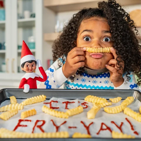 A girl holds a french fry as her elf watches in front of a tray scattered with fries, with "It's Fry-yay!" written in ketchup