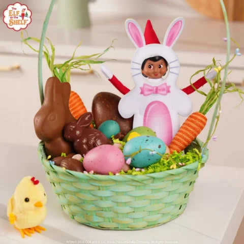 A girl elf is dressed in a bunny costume and is sitting in an Easter basket full of chocolate and colorful eggs
