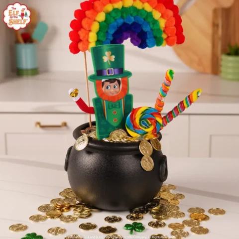 A boy Scout Elf is dressed in a leprechaun costume and is sitting in a pot of gold with a rainbow ovr his head