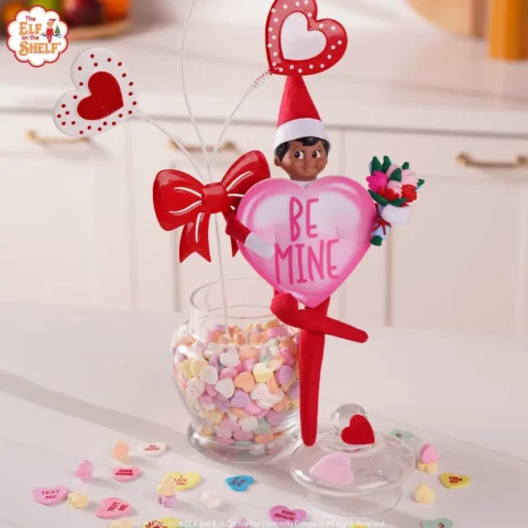A Scut Elf is dressed in a pink heart costume sitting on a jar of candy hearts