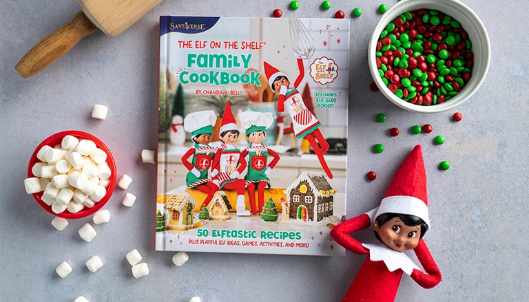 A Scout Elf posing beside chocolate candies, The Elf on the Shelf Family Cookbook, marshmallows and a rolling pin on a kitchen countertop.