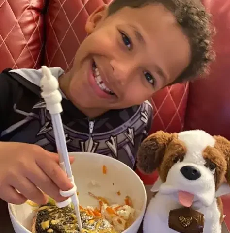 A young boy with curly brown hair eats noodles with chopstix while his Elf Pets Saint Bernard pup sits closeby
